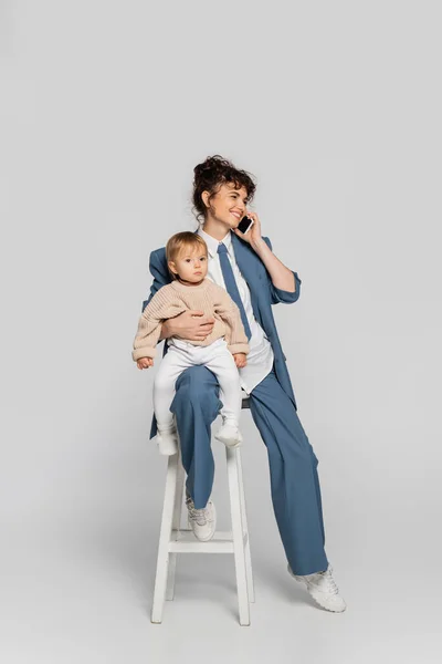 Happy businesswoman in blue suit talking on smartphone and sitting with toddler daughter on high chair on grey - foto de stock