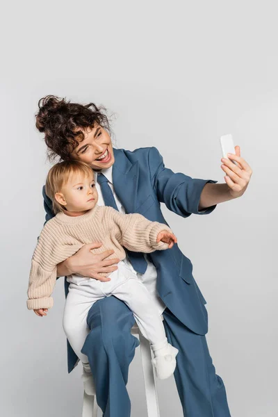 Happy businesswoman in blue suit taking selfie with toddler daughter on chair isolated on grey — Stockfoto