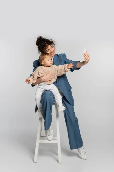 Cheerful businesswoman in blue suit taking selfie with toddler daughter on high chair on grey — Fotografia de Stock