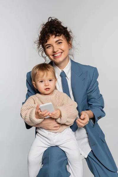 Happy businesswoman in blue suit sitting on chair while toddler girl holding smartphone isolated on grey — Photo de stock