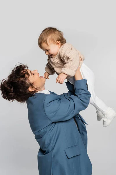 Happy businesswoman in blue suit holding cheerful toddler girl in sweater isolated on grey — Stockfoto