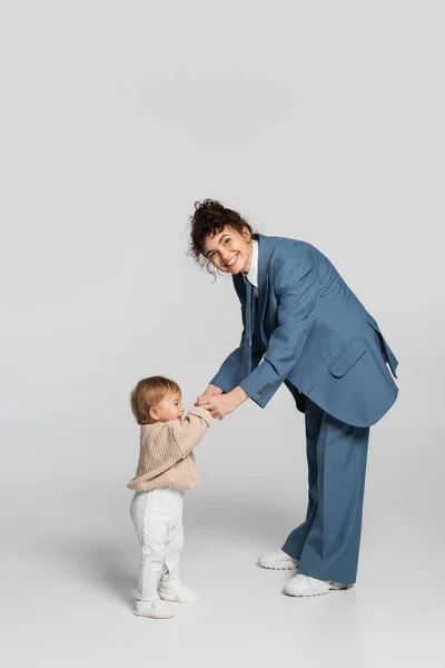 Full length of happy businesswoman in blue suit holding hands with baby girl in sweater on grey — Stock Photo