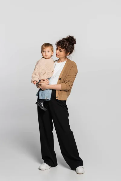 Full length of stylish mother in casual clothes holding in arms baby girl in sweater on grey - foto de stock