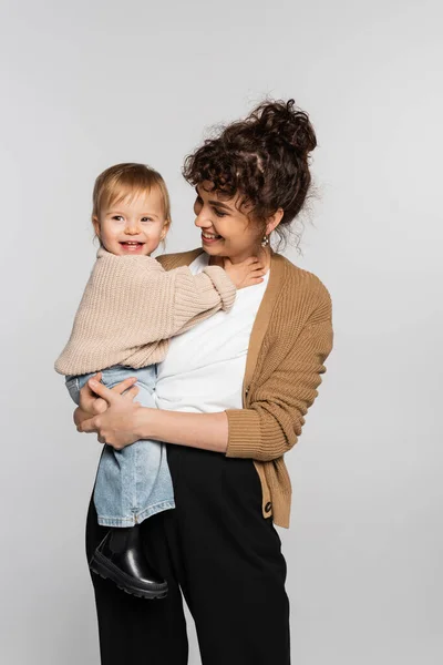 Happy mother in casual clothes holding in arms cheerful baby girl in sweater isolated on grey — Stock Photo