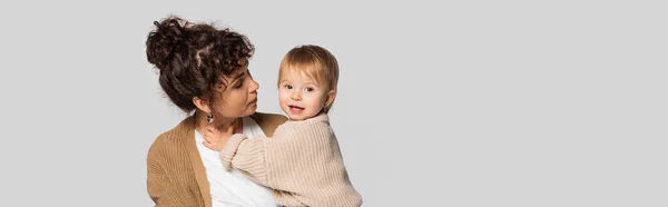 Stylish mother in casual clothes holding in arms cheerful baby girl in sweater isolated on grey, banner — Stock Photo