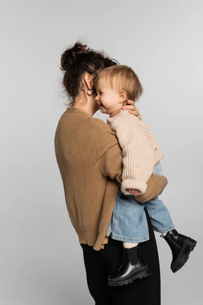 Curly mother in casual clothes holding in arms cheerful baby girl in sweater isolated on grey — Stock Photo