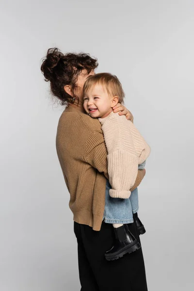 Curly mother in casual clothes holding in arms happy baby daughter in sweater isolated on grey — Stock Photo