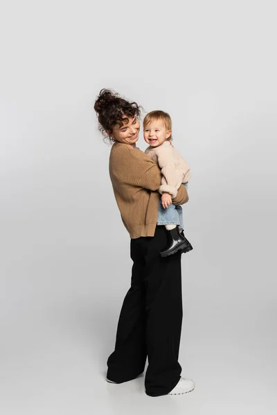 Longitud completa de madre feliz en ropa casual sosteniendo en brazos alegre bebé hija en suéter en gris - foto de stock