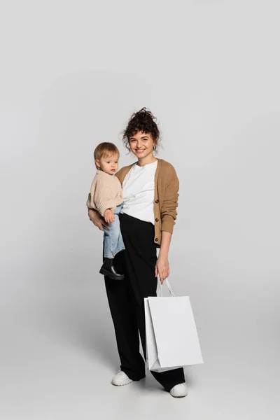 Full length of happy mother in casual clothes holding in arms cheerful baby daughter and shopping bag on grey — Photo de stock