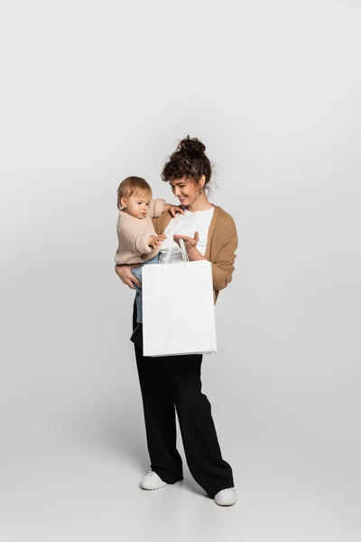 Full length of cheerful mother in casual clothes holding in arms daughter and shopping bag on grey — Stock Photo