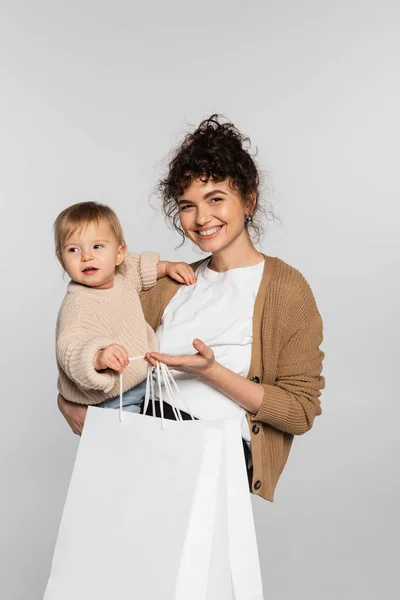 Cheerful mother in casual clothes holding in arms daughter and shopping bags isolated on grey — Photo de stock