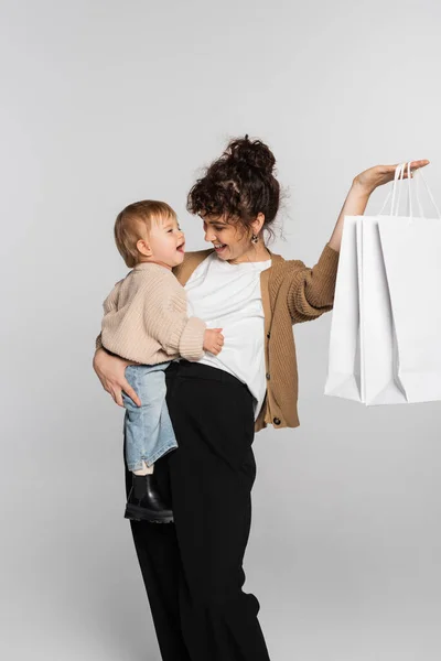 Happy mother in casual clothes holding in arms toddler daughter and shopping bags isolated on grey - foto de stock
