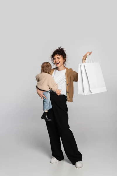 Full length of cheerful mother in casual clothes holding in arms toddler baby and shopping bags on grey — Stockfoto
