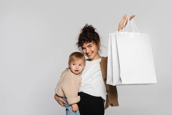 Smiling woman in casual clothes holding in arms toddler daughter and shopping bags isolated on grey — Photo de stock