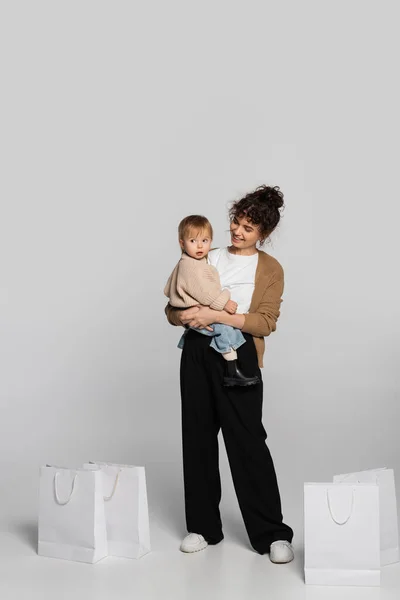 Full length of cheerful mother in casual clothes holding in arms toddler baby near shopping bags on grey - foto de stock