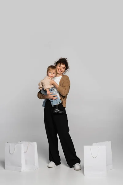 Full length of cheerful mother in casual clothes holding in arms toddler girl near shopping bags on grey - foto de stock