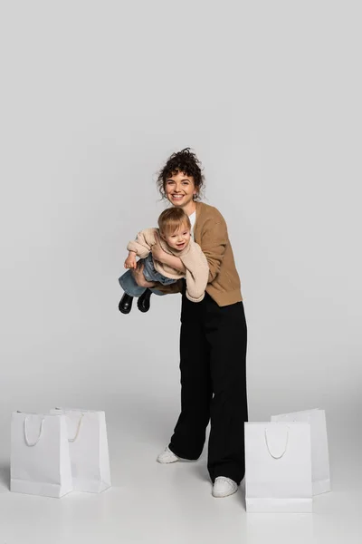 Full length of cheerful woman in casual clothes holding in arms toddler girl near shopping bags on grey — Stock Photo