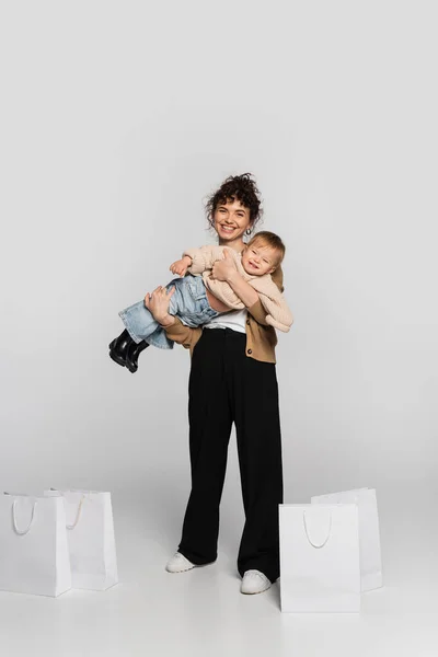Full length of smiling woman in casual clothes holding in arms toddler baby near shopping bags on grey — Photo de stock
