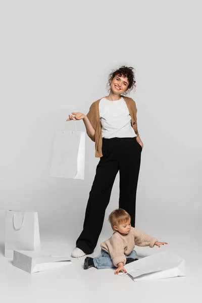 Full length of cheerful woman in casual clothes posing with hand in pocket near toddler daughter and shopping bags on grey - foto de stock