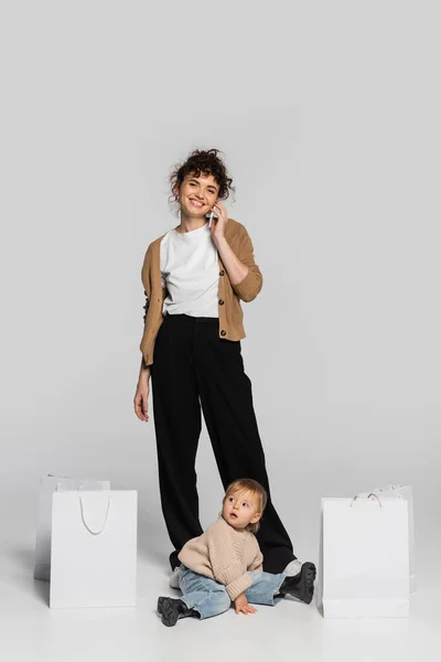 Full length of cheerful woman in casual clothes talking on smartphone near toddler daughter and shopping bags on grey — Stock Photo