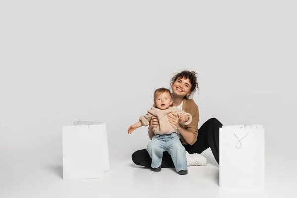 Full length of cheerful mother in casual clothes playing with toddler baby near shopping bags on grey — Stock Photo
