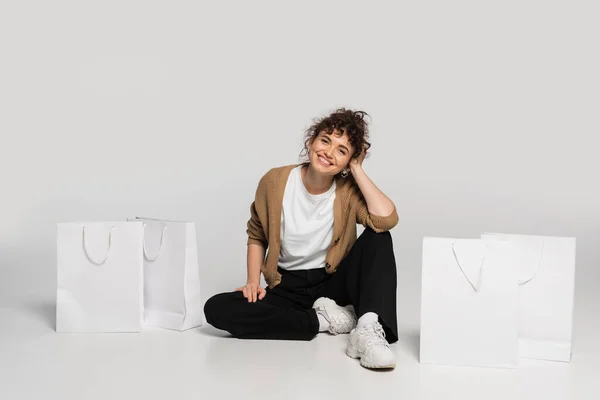 Full length of cheerful woman in casual clothes sitting near shopping bags on grey — Foto stock