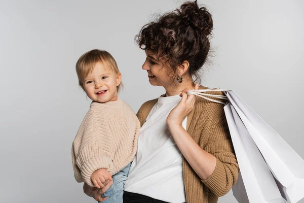 Mère heureuse en vêtements décontractés tenant dans les bras souriant tout-petit fille et sacs à provisions isolés sur gris — Photo de stock