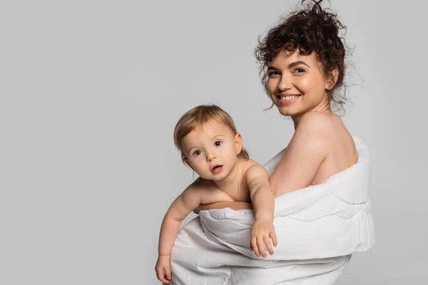 Joyful mother and toddler baby girl covered in duvet isolated on grey — Photo de stock