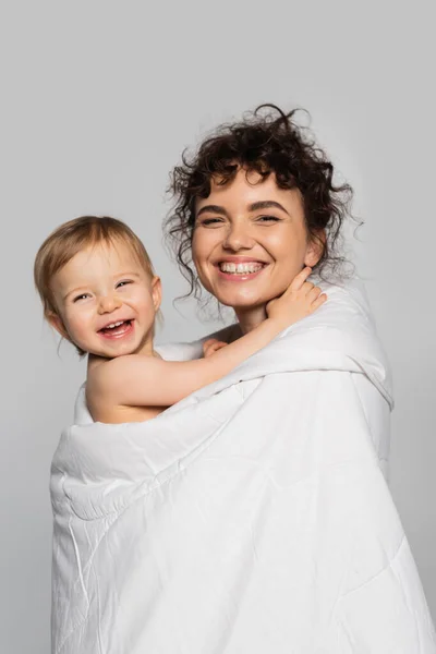Feliz madre y alegre hija niño cubierto de edredón aislado en gris - foto de stock
