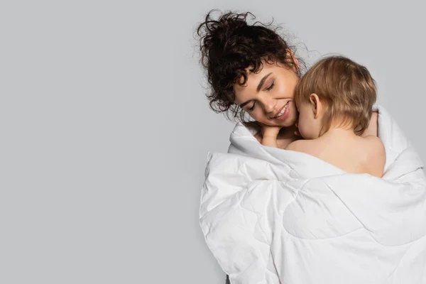 Happy mother with closed eyes hugging toddler daughter covered in duvet isolated on grey — Photo de stock