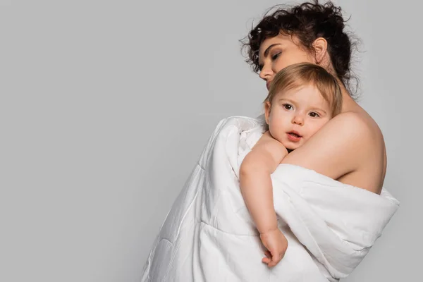 Curly mother hugging toddler daughter covered in white duvet isolated on grey — Stock Photo
