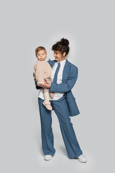 Full length of happy businesswoman in blue suit standing and holding toddler daughter on grey — Stock Photo