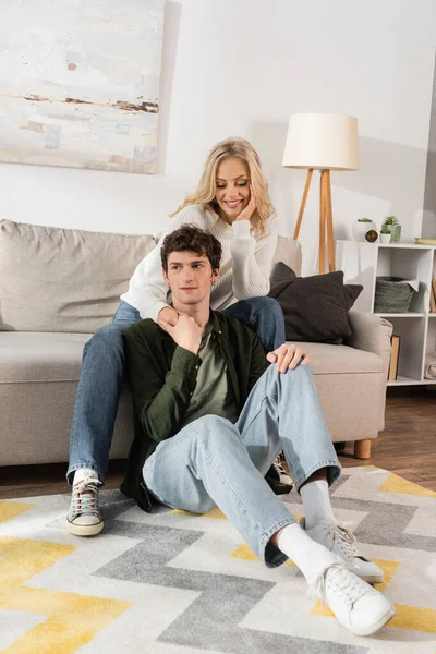 Full length of young blonde woman in white sweater hugging curly boyfriend in living room — Foto stock