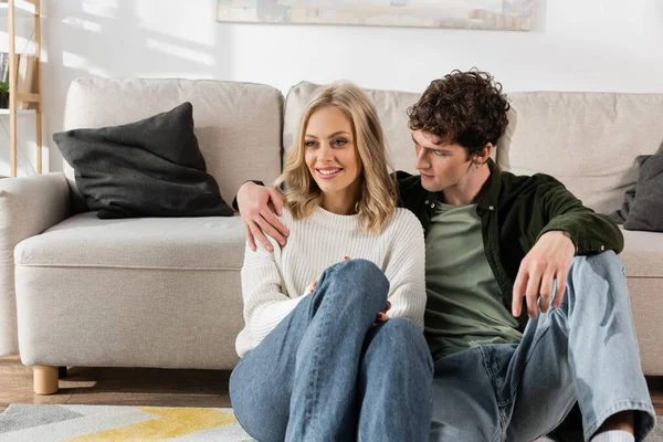 Curly man hugging pretty blonde woman sitting near sofa in living room — Foto stock