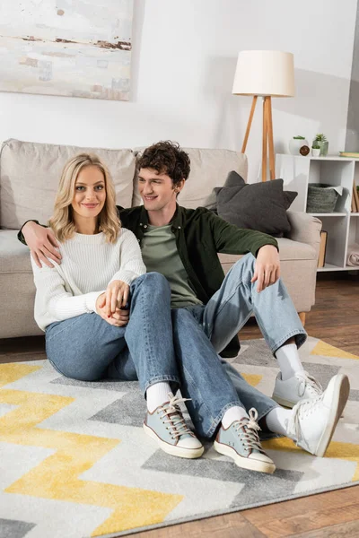 Full length of curly man hugging blonde woman sitting near sofa in living room — Foto stock