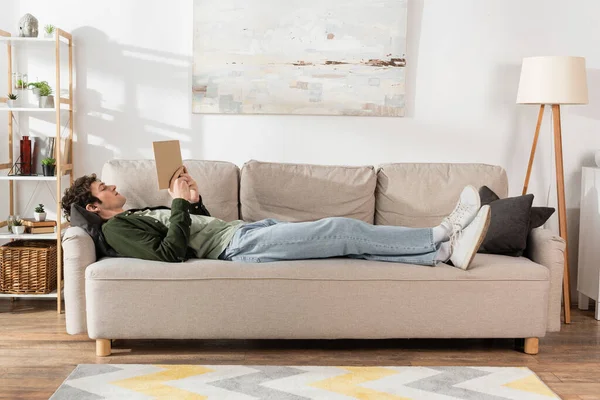 Full length of young man with curly hair reading book while lying on couch in living room — Stock Photo