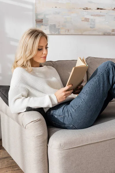 Young blonde woman in white sweater and jeans reading book in living room — Stock Photo