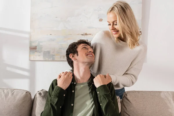 Young cheerful woman in white sweater hugging boyfriend in living room — стоковое фото