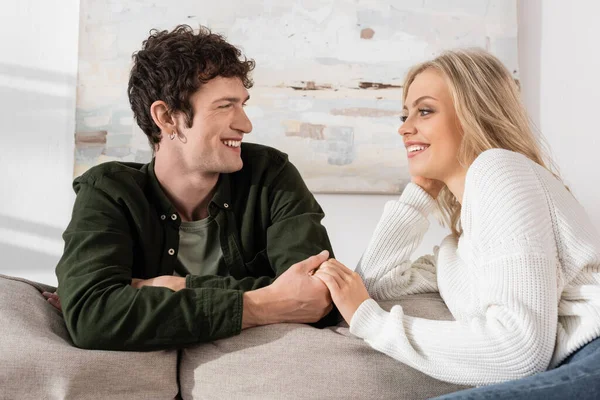 Cheerful young woman with blonde hair looking at curly boyfriend in living room — Foto stock