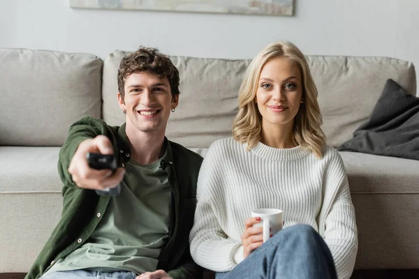 Cheerful man with curly hair clicking channels with remote controller near smiling girlfriend with cup of coffee — Stock Photo