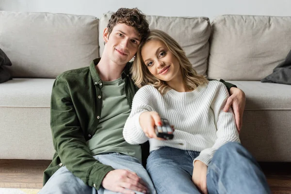 Happy blonde woman clicking channels with remote controller near boyfriend in living room — стоковое фото