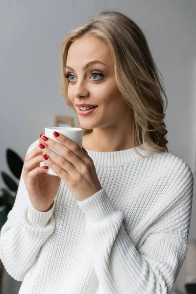 Young blonde woman in white sweater holding cup of tea and looking away at home — Stock Photo