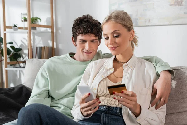 Cheerful woman holding credit card and smartphone while doing online shopping near boyfriend — стоковое фото