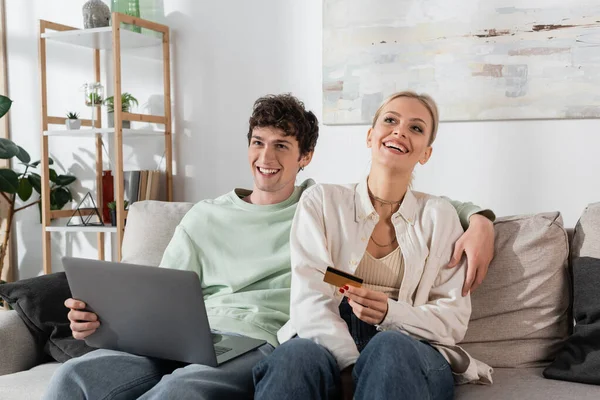 Cheerful woman holding credit card near boyfriend with laptop doing online shopping — Stock Photo