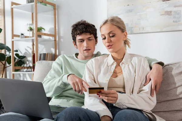 Curly man hugging blonde girlfriend and pointing at credit card near laptop — Stock Photo