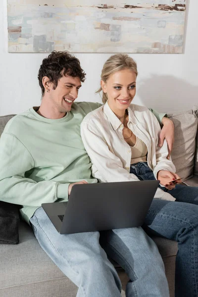 Cheerful woman holding credit card near boyfriend using laptop at home — Stock Photo