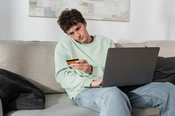 Curly young man holding credit card while talking on smartphone near laptop — Photo de stock