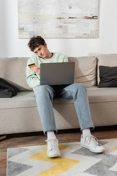 Curly young man holding credit card while making order and talking on smartphone near laptop — Stock Photo