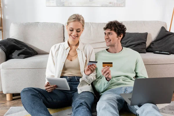 Cheerful man and woman holding gadgets and credit cards while doing online shopping — Stock Photo