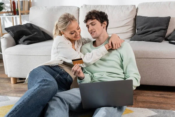Happy blonde woman hugging boyfriend holding credit card while using laptop — Stock Photo